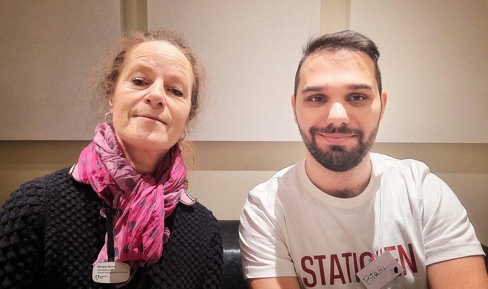 En vit kvinna med långt ihopsatt hår med en lila skarf runt halsen sitter bredvid en man med skägg, brunt kortklippt hår och en vit tshirt som det står Stationen på.