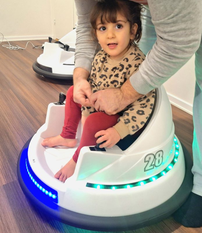 Treårig flicka sitter i en rund vit radiobil och får hjälp med att sätta på bältet.