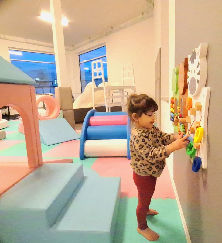 Treårig flicka i röda byxor och brunmönstrad tröja, brunt håri hästsvans står och leker med lekpanel, ibakgrunden mjeka lekmoduller.