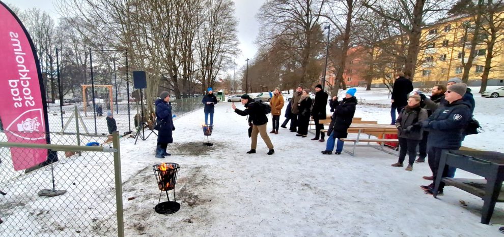 En grupp människor står och tittar på en kvinna som håller en mikrofon. I mitten står en man och fotograferar henne. Det är snö på marken och en liten eldstad närmast kameran.