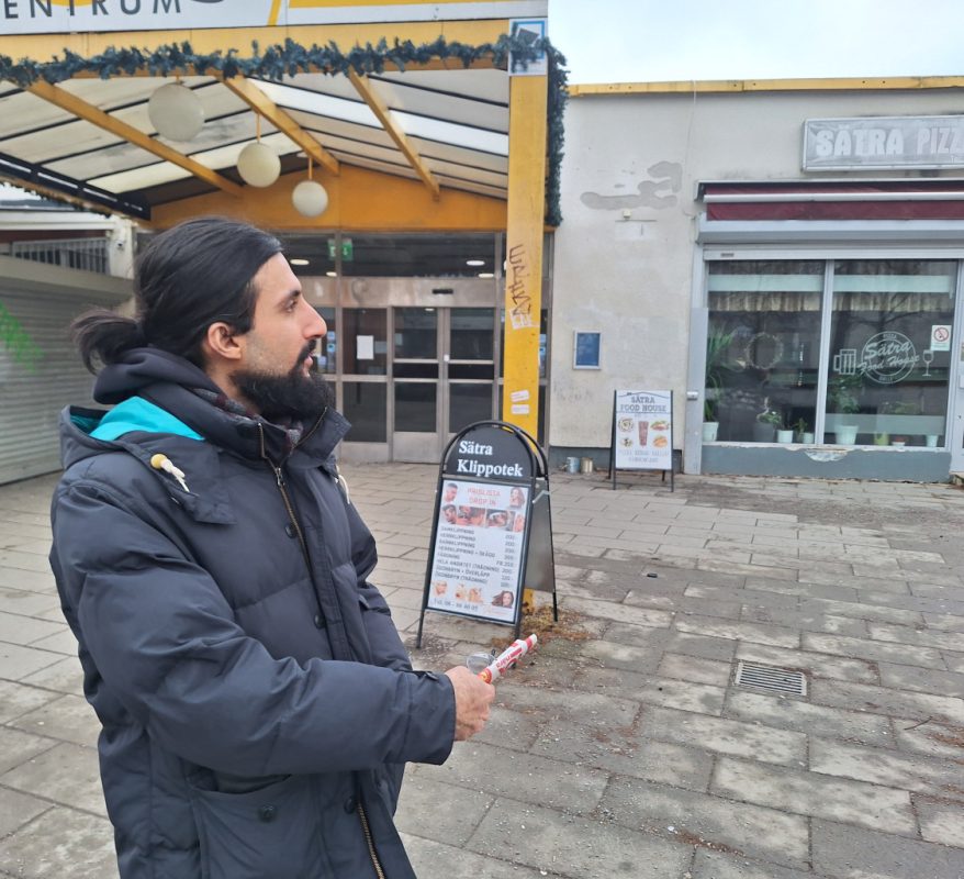 En man med svart hår i hästsvans och skägg, mörk täckjacka tittar i sidled. Han står framför en ingång till ett köpcentrum med gul inramning och vita väggar med flagnad målarfärg. Till höger en pizzeria med stora glasfönster.