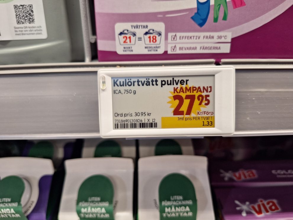 En vit rektangulär skylt. Med svart och röd text och en gul splash, som en sol med taggar i kanterna.