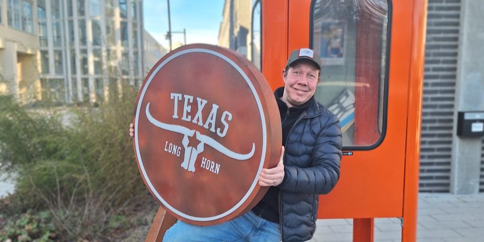 En vit man i mörk keps, blå täckjacka håller i en stor rund brun reklamskylt där det står Texas Long Horn på. Han står framför en orange telefonkiosk.