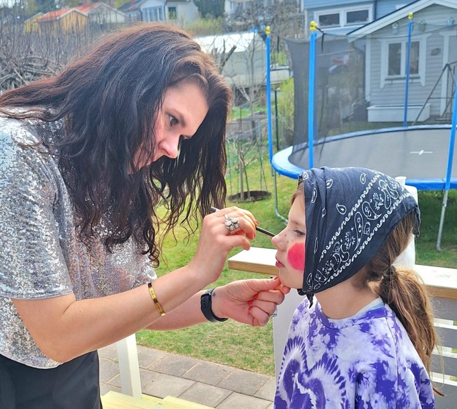 En vit kvinna med långt mörkt hår och silvrig blus står med en pensel och gör svarta prickar på en flicka med rödmålade kinder och huckle.
