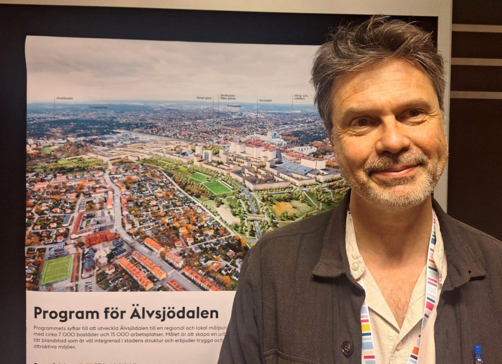 En medelåldersman i grått skägg mörkt kort hår. Ljus skjorta och brun kavaj framför en affisch som visar en skiss på en stadsdel.