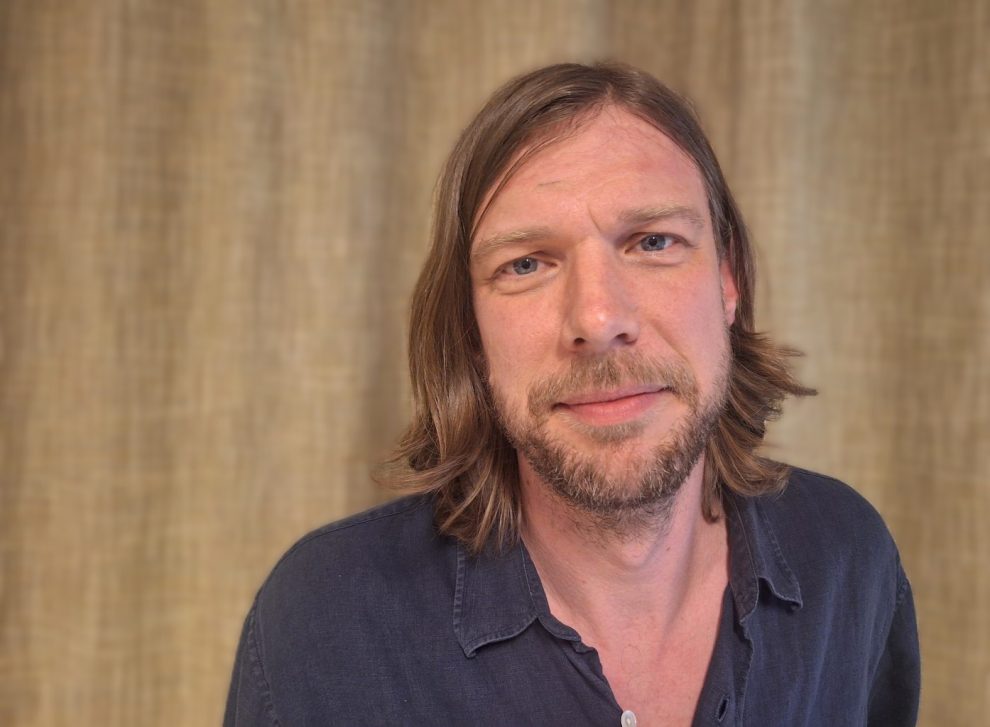 Vit man med axellångt hår och skägg, cendre och blå skjorta.