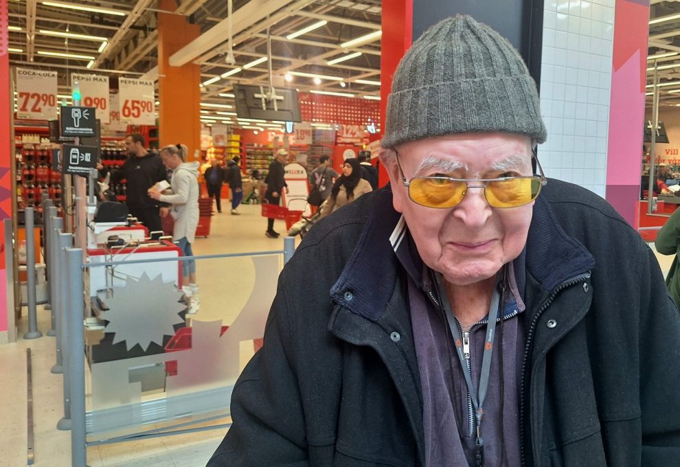Äldre man i grön stickad mösaa och gultonade glasögon, svart jacka.