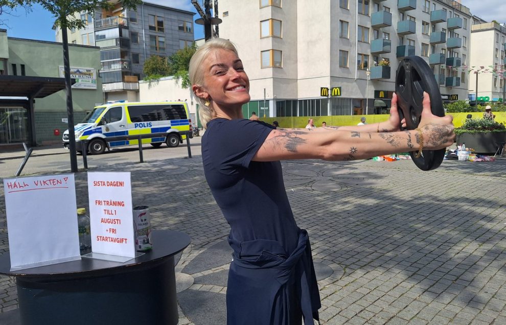 En vit kvinna med blont hår och mörkblå träningskläder såtr och håller i en rund vikt med armarna utsträckta.