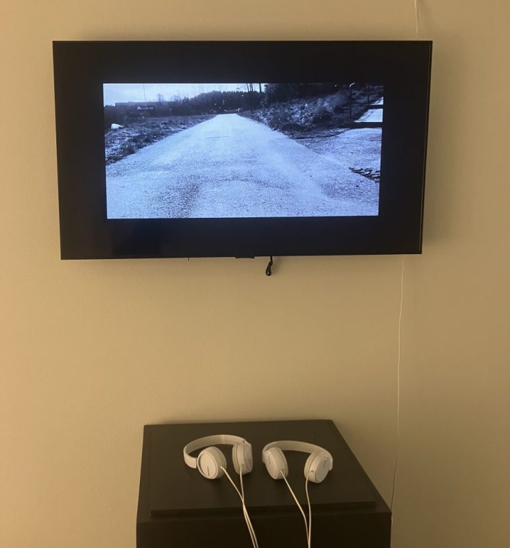 En tvskärm på en vit vägg. på ett litet bord ligger två vita hörlurar. På skären syns en grusväg i svartoch vitt