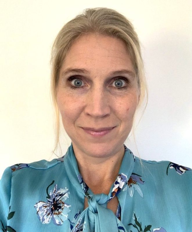 En vit kvinna med blont uppsatt hår och turkos blommig blu.