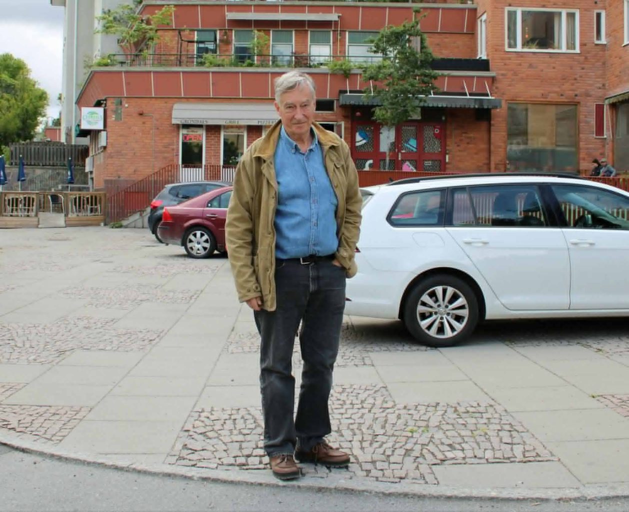 Äldre vit man med blå skjorta och beige jacka står vid en parkeringen med ett rött tegelhus i bakgrunden