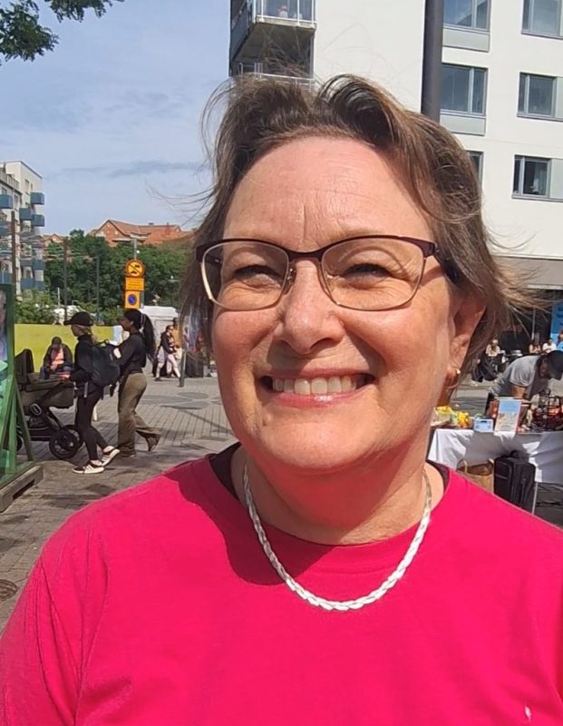 Vit kvinna med kort ljust hår, glasögon och en rosa t-shirt.