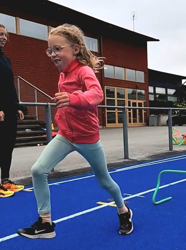 En sjuåring, blond vit flicka i glasögon, rosa träningsjacka och turkosa tights. Springer på en blå löparbana.