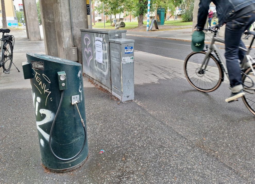 En mörkgrön rund, cirka en meter hög cykelpumpstation med svart lanag fäst vid sidan. I bakgrunden ett grått elskåp, bilväg och en cyklist som passerar.