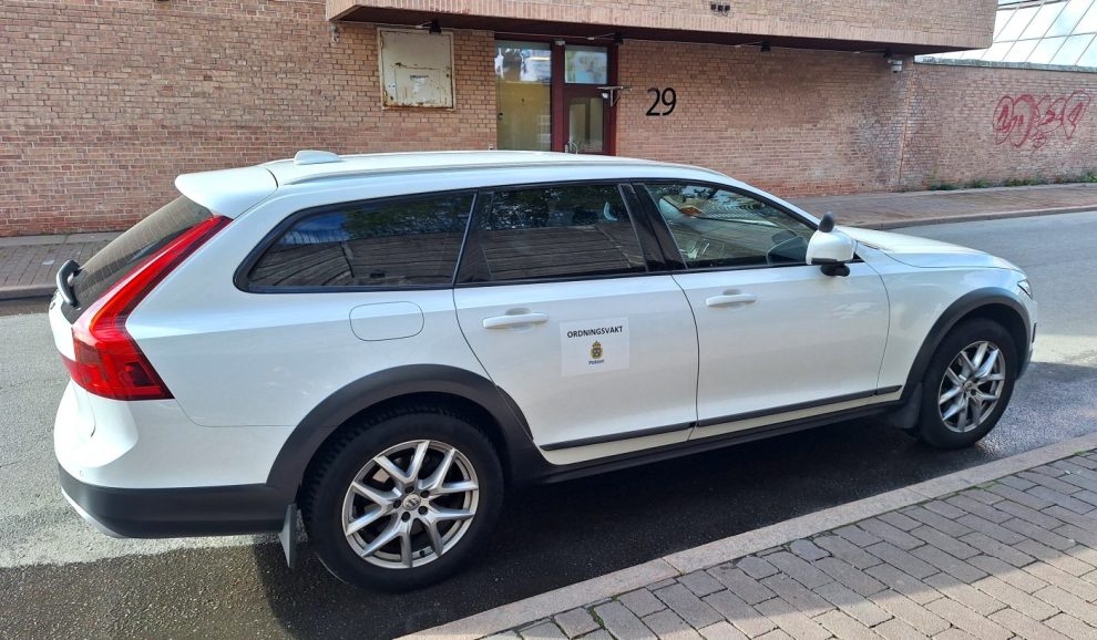 En vit bil står parkerad på en gata. I bakgrunden ett rött tegelhus