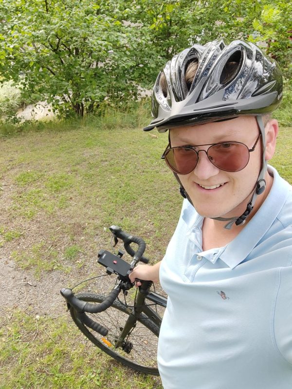 En vit man med svart cykelhjälm står och håller i en cykel. På marken gräs och buskar i bakgrunden. Han bär en ljusblå t-shirt.