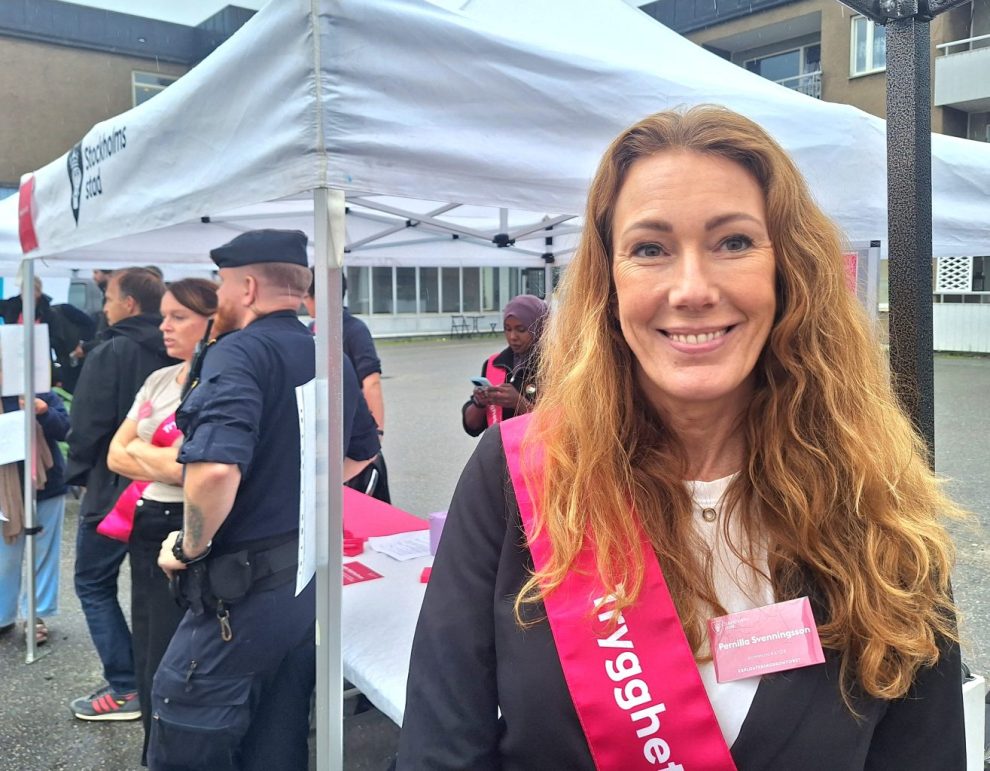 Vit kvinbna i längt rödblont hår, svat jacka med en rosa banderoll runt axeln.