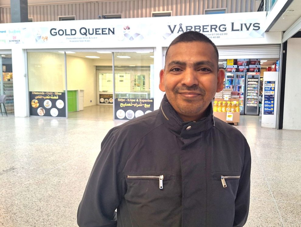 En svart man med svart jacka, kort hår ståri ett köpcentrum.