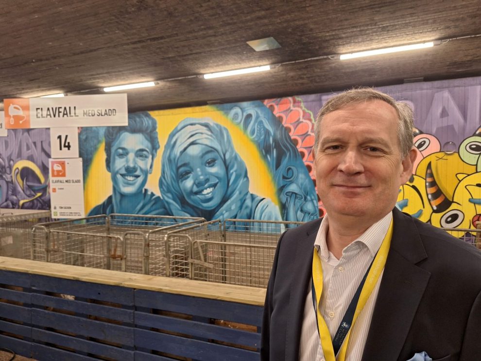 En vit man med kort grått hår framför en muralmålninge i turskosa och gula färger med två proträtt på två barn.