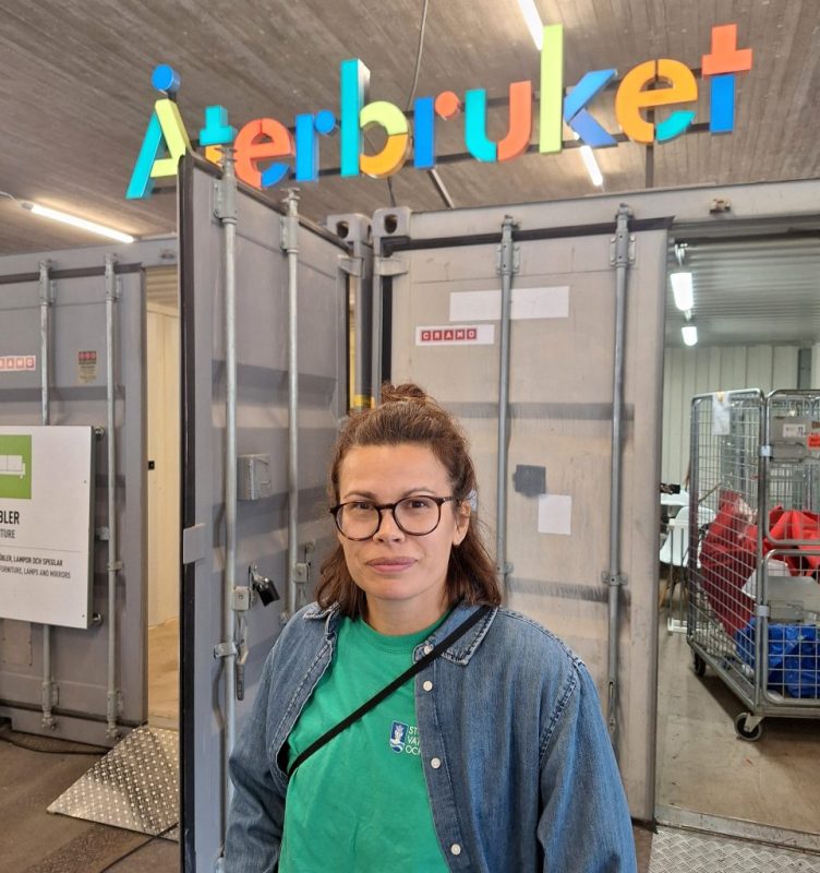 Vit kvinna i grön tshirt och blå jeansjacka, axellångt centrefärgat hår och glasögon. Står framför gråa containrar.