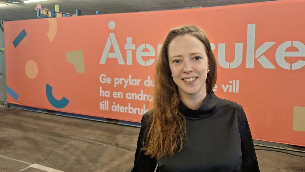 Vit kvinna i långt cendrefärgat hår och svart tröjda framför en rosa skylt som det står Återbruket på.