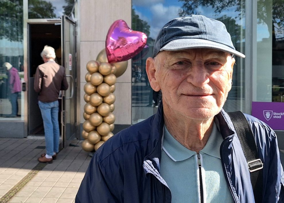En vit äldre man i blå keps och blå jacka. I bakgrunden en rosa ballong i from av ett hjärta.