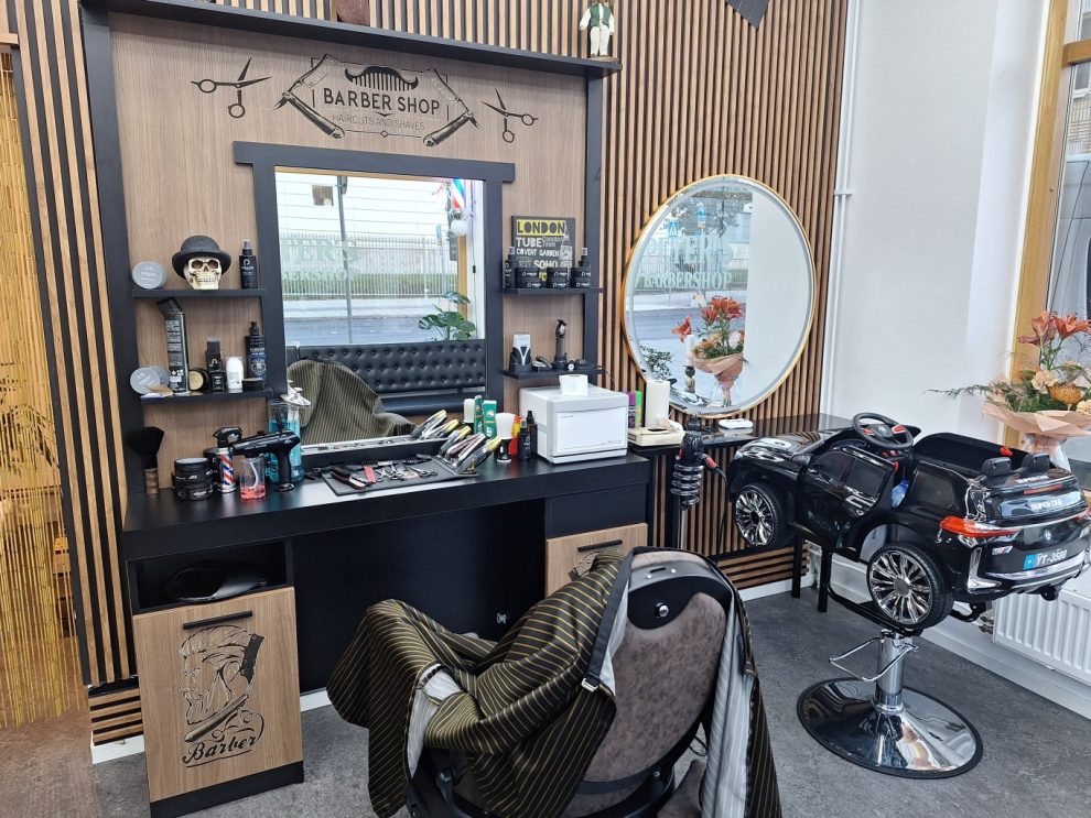 En frisörstol och bredvid en liten bil på ett stativ. En rund och fyrkantig spegel, klippverktyg och förkläde. Träpanel på väggarna.