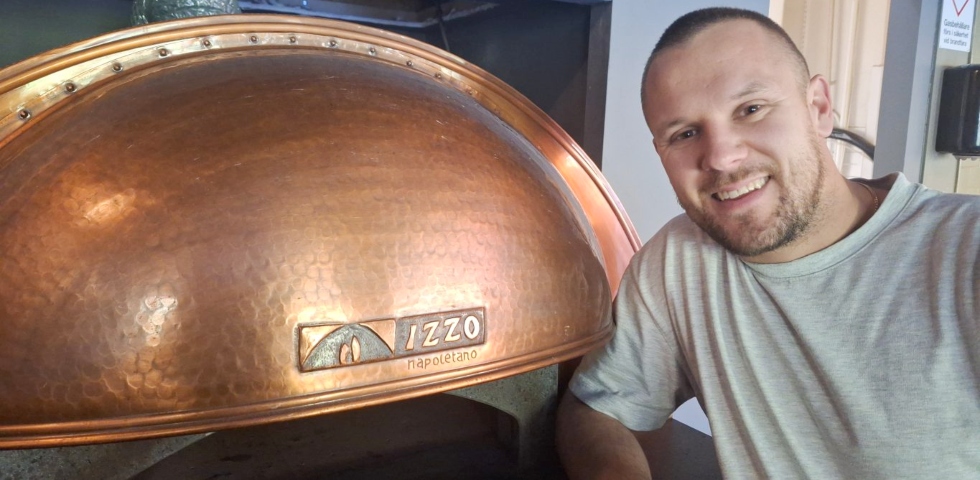 En vit man med snaggag hår och kort skäggstubb står lutad mot en pizzaung med kopparfärgat tak.