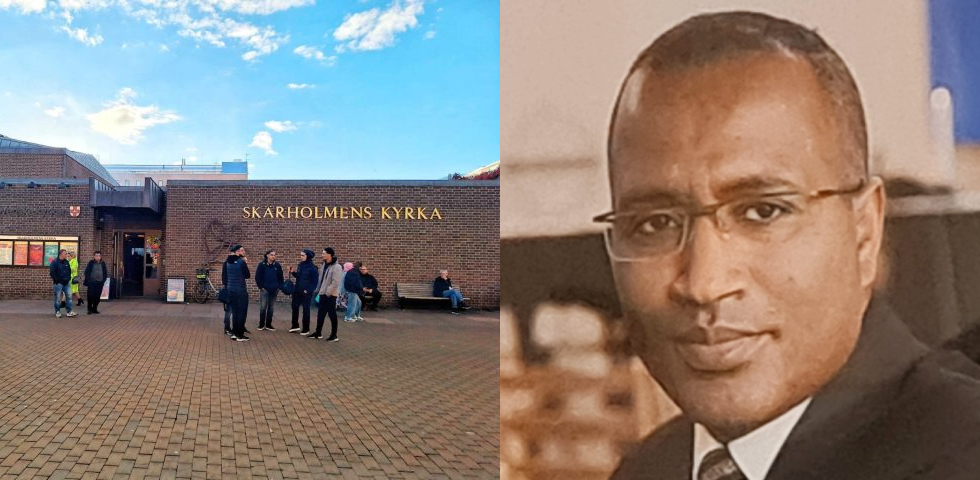 En svart med kallt huvud, glasögon, ljus kavaj och slips. Till vänster en låg, brun tegelbyggnad med människor utanför på ett torg