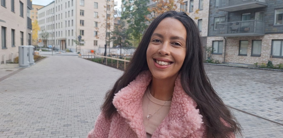 En brun kvinna med långt brunt hår och rosa fluffig jacka står på ett torg.