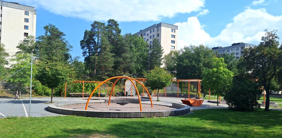 Enlekplats med klätterställningar i orangea färger. Gröna träd och höga hus i bakgruden.