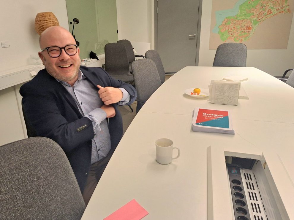 Vit man, kallt huvud, glasögon, mörk kavaj och ljus skjorta sitter i ett kontorslandskap.