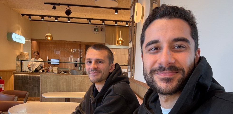 En vit man i kort hår och en icke vit barn i förgrunden med brunt hår och skägg och svarta kläder sitter på ett kafé.