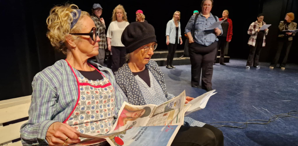 Två äldre kvinnor sitter på en vit bänk och läser tidningen. I bakgrunden syns skådepelare.