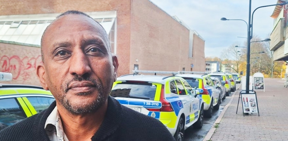 En svart man står med mörk jacka kort hår, på en gata med en rad polisbilar bakom sig.