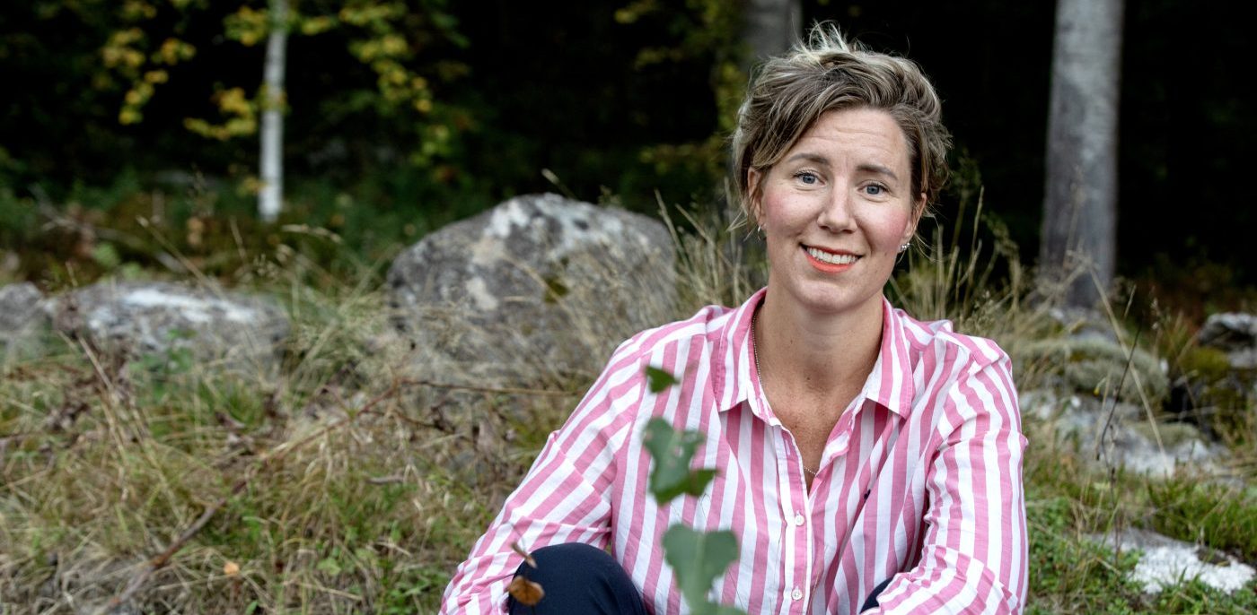 leende kvinna i blont hår och rödrandig skjorta, omgiven av träd och buskage i en höstmiljö