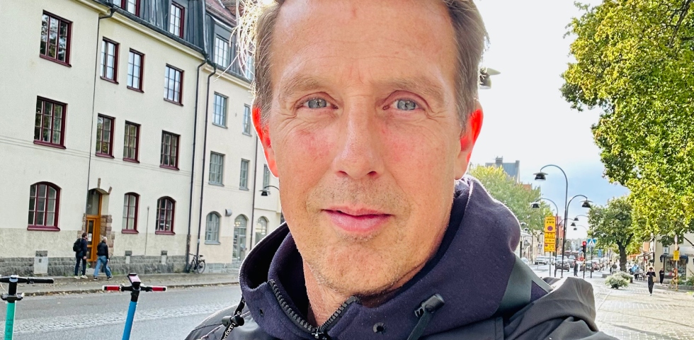 Vit man i kort hår , blåa ögon och mörkblå jacka. står framför ljusa husfasader.