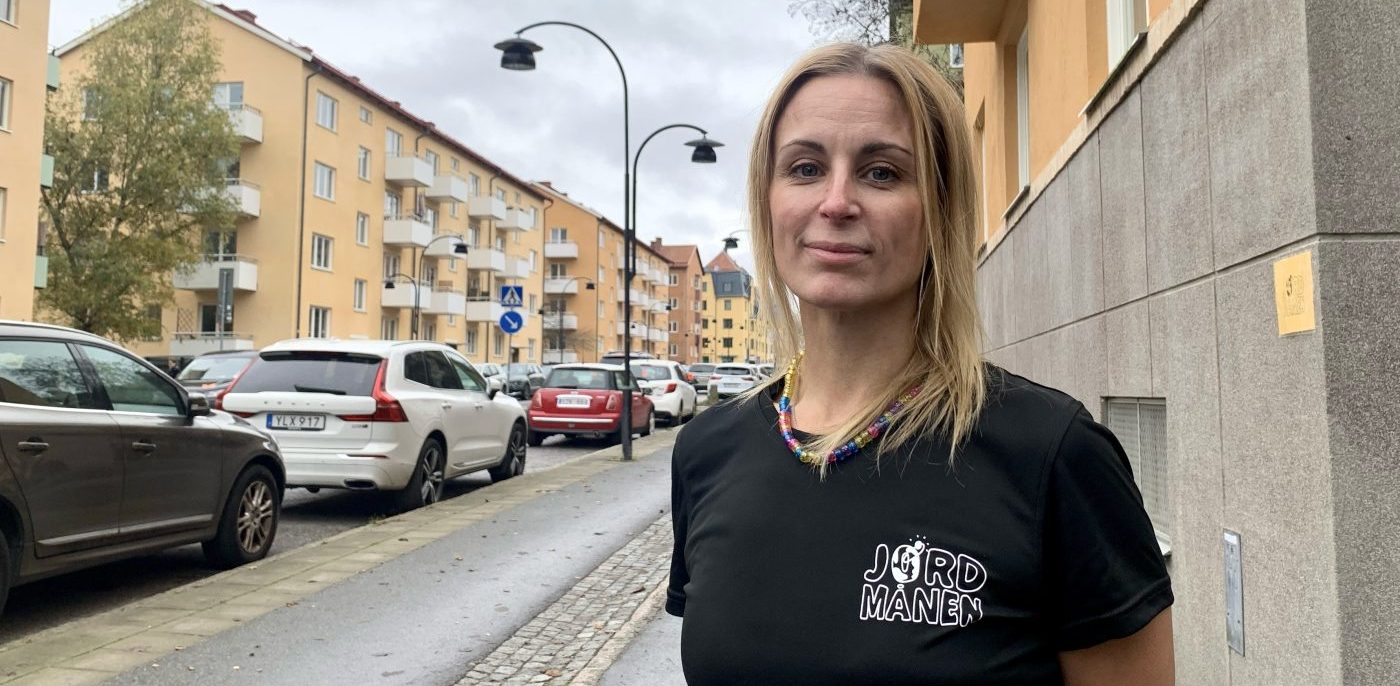 Blond kvinna med svart tröja framför en bildekorerad väg med låga lägenhetshus