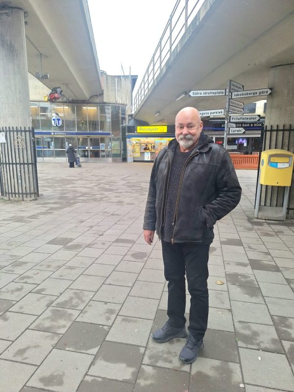 En äldre skallig man står i mörka kläder på ett torg framför ingången till en tbana och pressbyrå