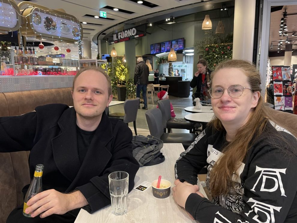 Till vänster: En vit man med kort hår och svart tröja sitter vid ett bord. Mittemot sitter en vit kvinna med långt cendréfrägat hår och glasögon med en tröja med vitt och svart tryck.