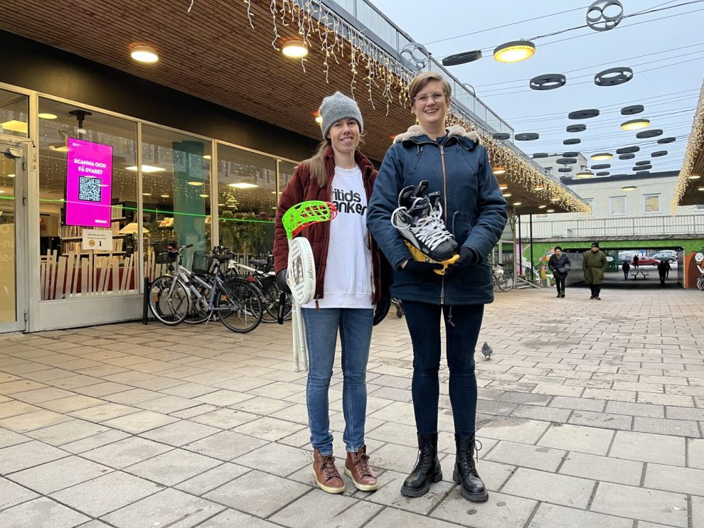 Två kvinnor står framför fritidsbankens planerade lokal, en med tennisracket och bandyklubbor i hand och en med skridskor i famnen.