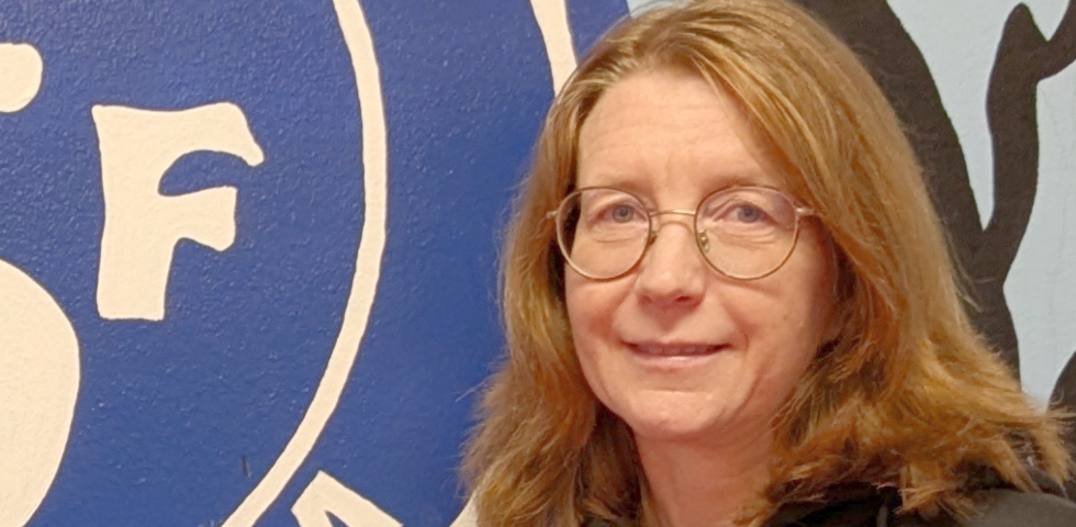 En medelålders vit kvinna i rödlätt, långt hår, glasögon och sn mörk träningsjacka.