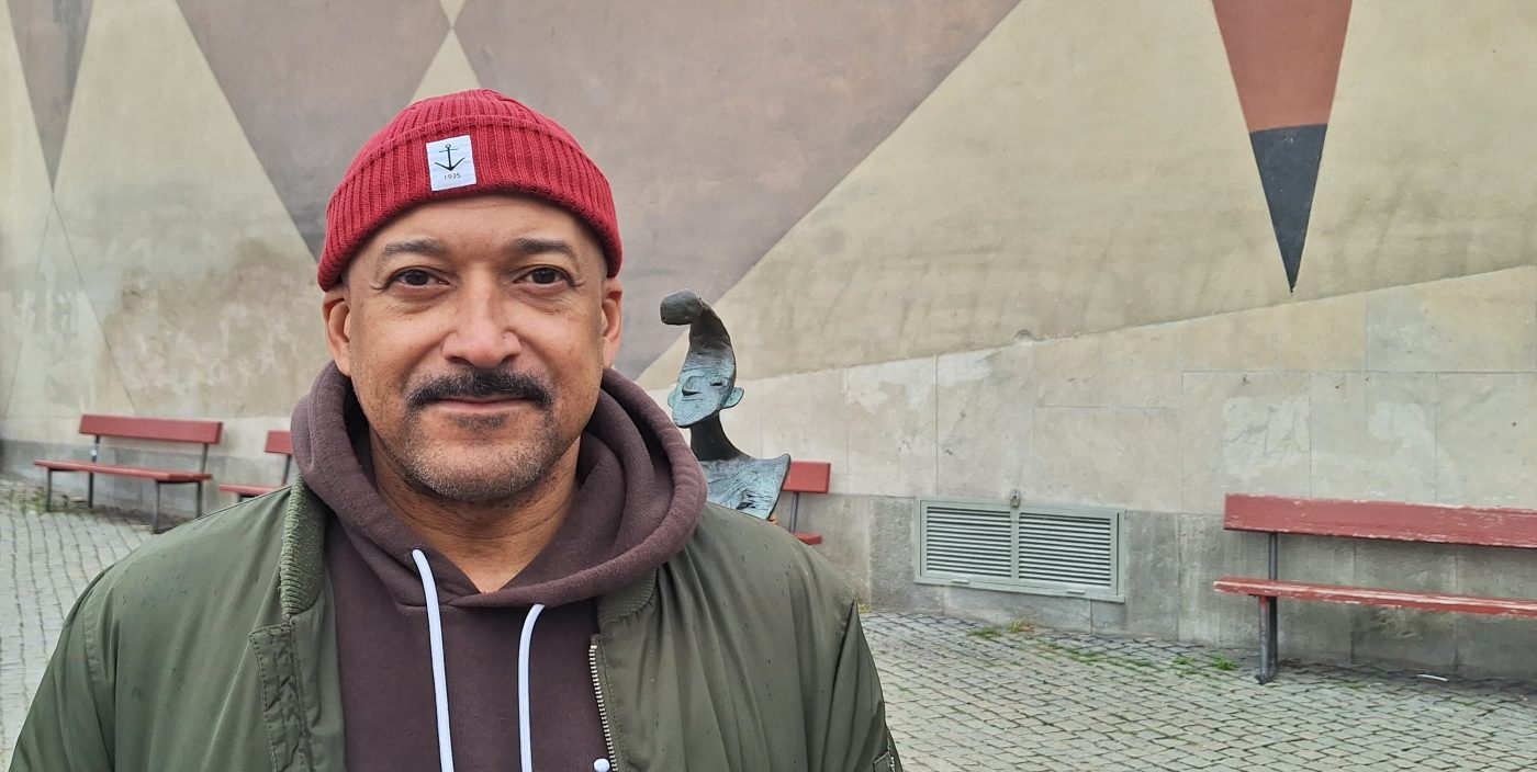 En svart man i röd mössa, brun hoody och grön jacka står framför en vägg målad i dova färger.