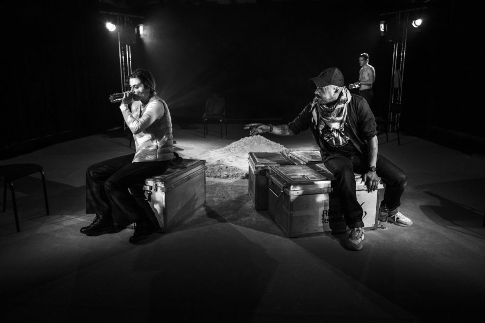 Ett svartvitt foto med två skådespelare som sitter på var sin låda i ett kallt scenrum med sand i bakgrunden.