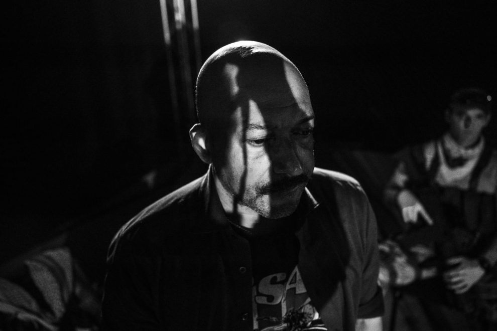 En svartvitbild, med en svart man utan hår, mörk t-shirt och står och tittar neråt med drmatiska skuggor över ansiktet.