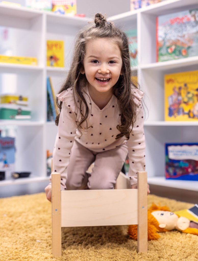 Smiling young girl with long hair crawls over a small wooden bed in a bright playroom with bookshelves in the background.