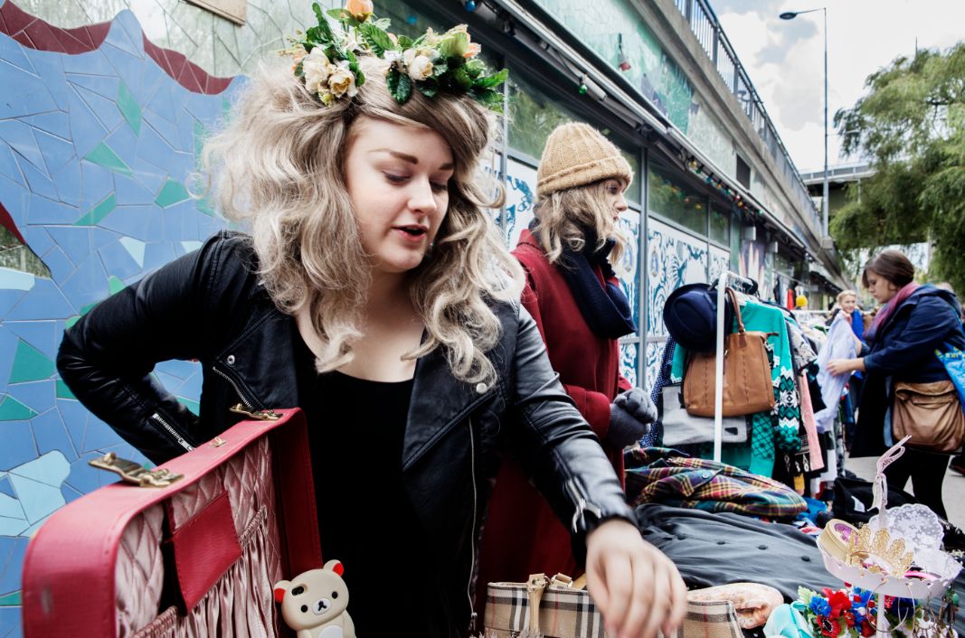 Woman with a flower crown browses a street market stall selling bags and accessories against a colorful mural.",