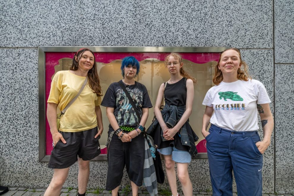 Four young adults posing together on a city sidewalk in front of a pink storefront window.