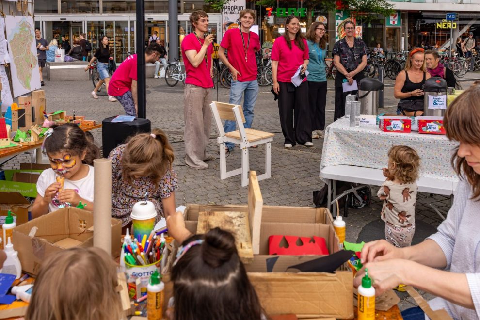 Children at a street craft table painting and crafting while volunteers in pink shirts stand nearby in a bustling urban square.
