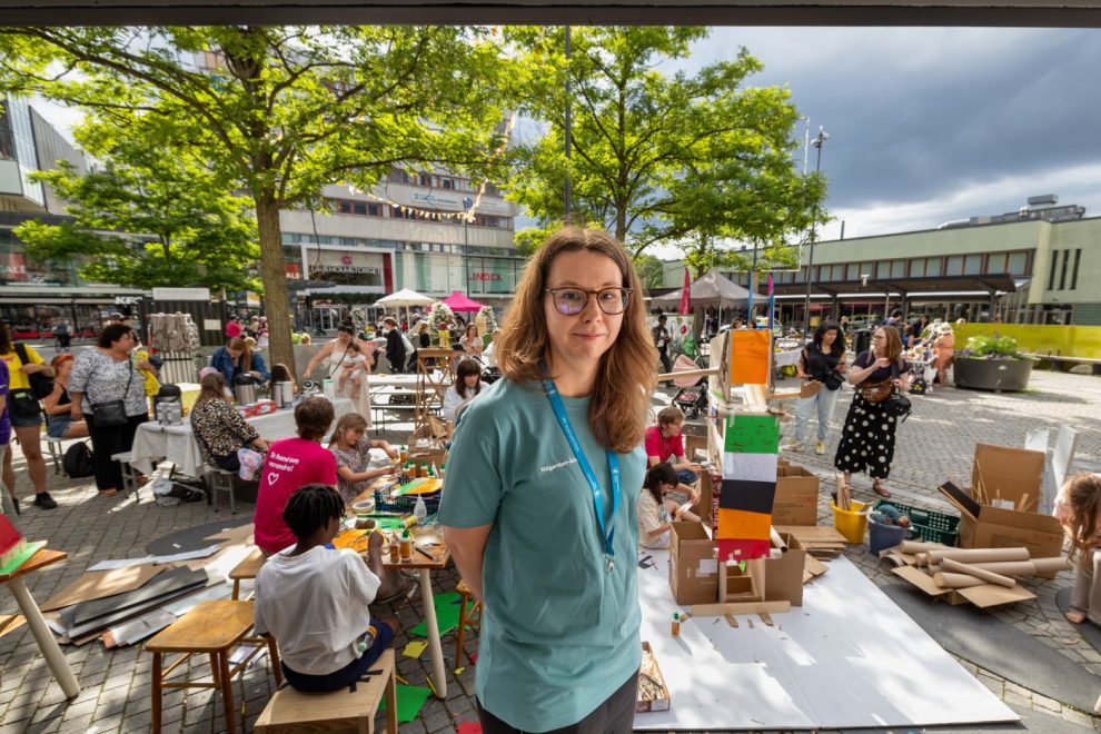 Woman with teal shirt and lanyard stands at a bustling outdoor craft market, colorful crafts and students at tables behind her.